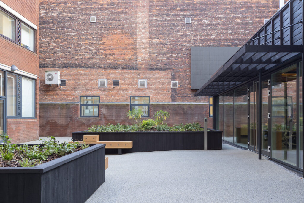 An outdoor courtyard area with a modern design, featuring raised black planters filled with greenery, wooden benches, and a gravel-like ground surface. The surrounding buildings have brick walls and windows, with one building displaying a mounted air conditioning unit. A glass wall on the right side reflects parts of the courtyard.