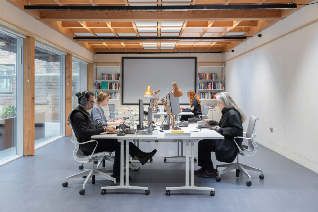 A modern office space with four people working at desks arranged in two rows facing each other. Large windows on the left allow natural light to fill the room. Bookshelves filled with books line the back wall, above which a large screen or whiteboard is mounted. Each desk has a computer monitor, desk lamp, and various office supplies. The ceiling features exposed wooden beams and recessed lighting.