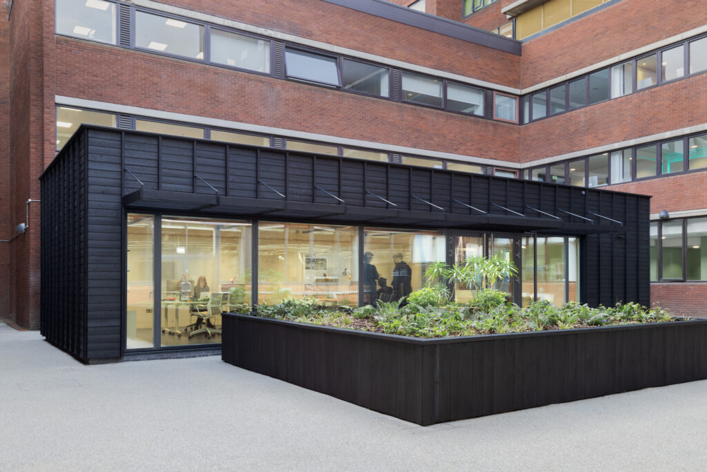 A modern building, the Work in Progress Pavilion, with a black exterior structure featuring large glass windows, through which an office space with people working is visible. In front of the structure is a rectangular planter box filled with various plants and greenery. The main building in the background is constructed of red brick and has multiple floors with windows.