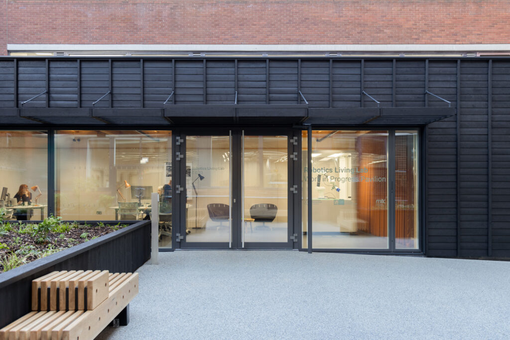 The exterior of the Work in Progress Pavillion with large glass windows and doors. The cladding on the building's exterior consists of horizontal black panels that are evenly spaced, giving the facade a sleek and modern look. Inside, people are visible working at desks with computers. Text on the glass reads 'Robotics Living Lab Work In Progress Pavilion.' In front of the building, there is a wooden bench and a planter filled with greenery.